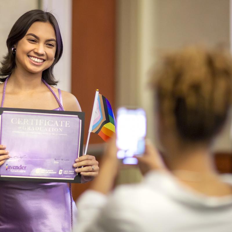 Graduating student with medium length dark hair. Wearing a purple dress holding a graduation certificate and LGBTQ+ Pride Flag.